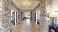 Located at 4203 River Birch Loop in High Point, NC, this single-floor community clubhouse delivers an impressive blend of rustic elegance and modern design. The grand entrance hall sets an immediate tone with dramatic stacked stone columns flanking both sides, herringbone-patterned wood-look tile flooring, and a striking geometric gold chandelier suspended beneath a tray ceiling with crown molding.

The open layout flows seamlessly into distinct functional zones. A welcoming lounge area centers around a floor-to-ceiling stacked stone fireplace, framed symmetrically by built-in white cabinetry and open shelving. Natural light fills the space through large windows dressed with soft linen curtains, while recessed lighting and ceiling fans maintain a bright, comfortable atmosphere throughout.

An office or leasing area features a workstation with ample desk space, bold artwork on the walls, and direct sightlines into the lounge, enhancing the open and collaborative feel of the space.

The clubhouse also includes a well-equipped bar and kitchenette, complete with a curved island with bar seating, white quartz countertops, white cabinetry, a stainless steel sink and appliances, and wall-mounted televisions — ideal for community gatherings and events. Consistent cool gray-blue wall tones and warm wood flooring tie the entire space together into a cohesive, inviting environment.