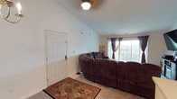 This single-floor residence offers a practical and comfortable layout across several well-defined spaces. The living room serves as the focal point of the home, distinguished by its vaulted ceilings that lend a sense of openness and height. A ceiling fan with a light fixture is centrally positioned overhead, while a decorative chandelier adds a touch of warmth near the entry. The front door opens directly into this space, which is anchored by soft carpeting, a patterned area rug, and a large sectional sofa. A window with drape treatments at the far end allows natural light to filter in and offers an outdoor view.

The kitchen follows a galley-style configuration with hardwood-style flooring, honey-toned wood cabinetry, white countertops, and white appliances including a range and refrigerator. An electrical panel is visibly mounted on the wall, and a peninsula counter separates the kitchen from the adjoining living space. Pendant lighting adds ambient illumination.

The bedroom features carpeted flooring, dark curtains on multiple windows, a ceiling fan, and ample room for a full furniture arrangement including a platform bed with built-in storage and a wall-mounted television.

A carpeted hallway connects the rooms and leads to a full bathroom finished with tile flooring, a vanity with wood cabinetry, and standard fixtures. An additional carpeted room with a window provides flexible use as a secondary bedroom or office.
