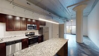 Located on Home Street in Cincinnati's vibrant 45202 zip code, this single-floor loft-style unit showcases an industrial-modern aesthetic throughout. The open-concept layout is defined by striking polished concrete floors with a marbled gray and white finish, complementing the exposed ductwork and structural columns that reinforce the space's urban character.

The kitchen is well-appointed with rich dark-walnut cabinetry, granite countertops, and a full suite of stainless steel appliances, including a range, built-in microwave, refrigerator, and dishwasher. A generous island provides additional prep and seating space, anchoring the kitchen as a central hub of the unit.

The living area extends toward a sliding glass door that opens to a private outdoor area, inviting natural light deep into the interior. Ceiling fans and track lighting provide practical illumination, while the tall ceilings with visible infrastructure lend an airy, loft-like atmosphere.

Additional rooms include a versatile enclosed space well-suited for a bedroom or home office, featuring the same polished concrete flooring. The bathroom is clean and functional, equipped with a full tub-shower combination in a bright, all-white finish. The overall ambiance is modern and industrial, ideal for urban living.