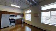 This single-floor space showcases an industrial loft aesthetic defined by exposed concrete ceilings with visible ductwork and polished concrete floors that reflect natural light throughout the open layout. The kitchen is positioned along one wall and features dark wood cabinetry, granite-style countertops, a stainless steel appliance package including a gas range, built-in microwave, and dishwasher, as well as a double-basin sink beneath upper-mounted windows. Natural light is a prominent feature, with multiple windows fitted with horizontal blinds positioned at varying heights along the walls, allowing light to flow in from multiple directions while maintaining privacy. The elevated window placement enhances the sense of height, contributing to the open, airy atmosphere characteristic of loft-style spaces. The main living area is open and flexible, offering generous space for a variety of furniture arrangements. A baseboard heating unit runs along the lower wall, and a wall-mounted utility panel is also visible. The overall ambiance is contemporary and urban, blending functional design with raw architectural elements.