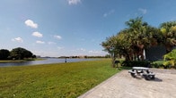 This single-story property offers a notable outdoor amenity in the form of a well-maintained waterfront green space. The exterior grounds are a standout feature, showcasing an expansive stretch of manicured lawn that leads directly to the edge of a calm, open body of water. The waterway provides broad, unobstructed views toward distant multi-story buildings on the far shoreline, set against a wide Florida sky. Mature palm trees and a flowering crepe myrtle tree add natural beauty and shade to the outdoor area. A concrete patio serves as a functional gathering point, equipped with a picnic table and a charcoal grill, making it well-suited for outdoor dining and recreation. The overall ambiance of the outdoor space is relaxed and scenic, benefiting from abundant natural light and open sky. The interior images provided are too dark to yield reliable detail; however, the property is laid out on a single floor. The combination of direct waterfront access, tropical landscaping, and dedicated outdoor amenities gives this space a distinctly leisure-oriented character consistent with a warm-weather, waterside lifestyle.