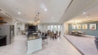 Located at 10300 Katy Freeway in Houston, Texas, this thoughtfully designed community amenity space offers a generous, single-floor layout ideal for socializing and gathering. The main open area features large-format white tile flooring and is bathed in natural light from floor-to-ceiling French doors that open onto a balcony with outdoor views. A sleek, dark-countertop island with bar seating anchors the central space, complemented by geometric brass pendant lighting overhead. Multiple dining configurations accommodate both intimate and larger group settings. The lounge area is anchored by a dramatic dark tile accent wall housing a built-in fireplace and flat-screen TV, flanked by open shelving. An industrial-style chandelier adds a modern edge to this comfortable seating zone. A secondary lounge area features plush carpeting, soft swivel chairs, and a teal accent wall with a wall-mounted display screen, creating a more relaxed retreat within the space. A private meeting room with carpeted floors, a sputnik chandelier, teal accent wall, and vibrant Western-themed artwork provides a focused setting for smaller gatherings or work sessions. Throughout, the design blends contemporary finishes with warm, inviting touches for a polished communal environment.