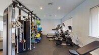 Located at 1070 Greenwall Pl in Frederick, Maryland, this fitness center offers a compact yet comprehensively outfitted exercise space designed to support a full range of workout routines. The room features rubber flooring throughout, providing a durable and slip-resistant surface ideal for high-activity use. Recessed ceiling lighting keeps the space bright and well-illuminated, while large windows fitted with vertical blinds allow natural light to filter in, contributing to an open and energizing atmosphere.

The cardio zone is equipped with multiple treadmills, an elliptical trainer, and a stationary recumbent bike, each outfitted with individual monitors for workout tracking. A wall-mounted flat-screen TV is positioned for easy viewing during exercise. On the strength training side, the facility includes a multi-functional cable and weight machine, a rack stocked with dumbbells of varying weights, and an adjustable workout bench. Stability balls and medicine balls in assorted sizes round out the functional fitness offerings.

A glass-paneled partition wall with a framed door separates one section of the space, adding a modern architectural touch while maintaining visual openness. A wall-mounted water fountain provides convenient hydration access. Clean white walls and a neutral color palette create a focused, distraction-free environment suited for daily fitness routines.