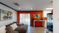 This single-floor commercial space blends bold design with practical functionality, creating an inviting and energetic environment. The main lounge area immediately sets the tone with a striking orange accent wall, wood-look flooring, and recessed lighting throughout. A large sliding glass door with vertical blinds fills the space with natural light while offering a view of the outdoor area beyond. A comfortable sectional sofa and a sleek white work console make the lounge equally suited for relaxation or productivity. Colorful artwork adorns the walls, reinforcing the space's vibrant aesthetic. Adjacent to the lounge, a private office features a dark wood executive desk, guest seating, and tasteful décor, providing a professional setting for meetings or focused work. Additional private rooms open off the main corridor, one of which is furnished with bold red accent chairs, a patterned area rug, and framed artwork — ideal as a consultation or meeting room. The hallway connects these spaces seamlessly with consistent trim detailing and dark-framed doors. The kitchenette is well-appointed with white shaker cabinets, a undermount sink, and a retro-style black refrigerator, complemented by whimsical artwork and natural light from a skylight above. Throughout the space, light wood-look flooring, dark crown molding, and curated décor create a cohesive, modern, and welcoming atmosphere.