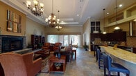 This single-floor community clubhouse offers a sophisticated and welcoming environment designed for both relaxation and socializing. The grand main lounge serves as the heart of the space, featuring soaring tray ceilings adorned with ornate wrought-iron chandeliers, a stone-surround fireplace, and generous natural light streaming through large French doors and arched windows. Slate tile flooring flows throughout the common areas, complemented by rich wood furnishings, plush seating arrangements, and decorative area rugs that anchor each gathering zone. The full kitchen is outfitted with dark espresso cabinetry, a mosaic tile backsplash, stainless steel appliances, and a curved granite-topped island with bar seating beneath elegant pendant lighting. A dedicated billiards room provides entertainment with a full-size pool table, Tiffany-style overhead lamp, wall-mounted television, and warm earth-toned walls. A separate media lounge features a stone-accented arched alcove housing a large flat-screen television, with curved sectional seating for comfortable viewing. A secondary dining and game area offers bistro-height tables with views to an outdoor covered patio. An art gallery-style hallway with track lighting and curated framed artwork connects the various areas, reinforcing the refined aesthetic carried throughout the entire space.