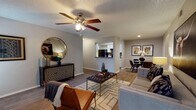 This contemporary living space showcases an open floor plan that seamlessly integrates multiple functional areas. The room features warm hardwood flooring and a textured ceiling adorned with a five-blade ceiling fan. A comfortable seating area is anchored by a plush gray sofa and a leather accent chair, centered around a tufted ottoman and geometric area rug. The space is further enhanced by a striking geometric-patterned sideboard, circular mirror, and curated wall art. A pass-through window offers a view into the kitchen, while a connected dining area is visible, emphasizing the flow between spaces. The neutral color palette and modern furnishings create a sophisticated and inviting atmosphere.
