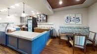 This single-floor space is thoughtfully designed as a welcoming community gathering area, combining functional service zones with comfortable social spaces. At its core is a well-appointed service counter featuring bold navy cabinetry topped with light granite-style countertops, a glass display case, and glass-front upper cabinets that provide organized storage for supplies and accessories. Pendant lighting and recessed ceiling fixtures illuminate the counter area effectively.

Adjacent to the service counter, a compact preparation area includes a sink, refrigeration unit, and additional cabinetry finished in a warm wood tone, complemented by a distinctive wood-paneled ceiling detail. A cozy built-in booth with a floral upholstered banquette offers an intimate dining spot nearby.

The main gathering area features rich hardwood flooring and is furnished with multiple round dining tables surrounded by cushioned wood chairs, creating a casual yet inviting atmosphere. A wall-mounted flat-screen television serves as a focal point in the lounge area. Large windows dressed with decorative valances overlook a covered outdoor patio, allowing natural light to fill the interior while providing pleasant views of the exterior seating area. The overall ambiance is warm, comfortable, and community-oriented.
