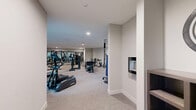 Located at 1850 Glenview Rd in Glenview, Illinois, this fitness center offers a spacious, single-floor exercise room designed to support a full range of workout routines. The room is finished with neutral gray walls and plush carpeted flooring, creating a clean and comfortable environment conducive to exercise.

The space is generously appointed with a diverse selection of equipment. Cardio offerings include multiple True-brand treadmills, elliptical trainers, recumbent bikes, and a stair climber, all positioned near large windows that flood the area with natural light and provide views of the surrounding exterior. For strength training, the room features a multi-station cable machine, a leg press, adjustable benches, and a dedicated free-weight area with a full dumbbell rack. A wall-mounted flat-screen television and full-length mirrors enhance both the functionality and the sense of openness within the space.

Recessed lighting throughout the ceiling ensures the room is well-lit during all hours, while a separate utility room door is accessible from within the exercise area. The layout is thoughtfully organized to allow ample circulation between equipment, making the space practical for multiple users simultaneously. Overall, this fitness center delivers a professional, amenity-rich environment suited for a broad spectrum of fitness activities.
