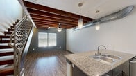 Located at 716 E Hickory St in Denton, TX, this two-story loft blends industrial design with modern finishes across two well-utilized levels. The ground floor centers around an open-concept kitchen and living area, where rich hardwood-style flooring and striking exposed wood ceiling beams set a warm, urban tone. The kitchen is well-appointed with dark cabinetry, granite countertops, a peninsula with a double stainless steel sink, black stainless appliances including a range and microwave, and pendant lighting overhead. Exposed ductwork runs along the ceiling, reinforcing the industrial character of the space. A ceiling fan adds comfort to the living area, which benefits from natural light through a large window with blinds. A wood-and-metal staircase with sleek horizontal metal railings connects the floors, serving as a architectural focal point in its own right. The upper level features plush carpeting, a ceiling fan, and views to the outside through additional windows. A dedicated closet provides storage on this level. The bathroom is finished with floor-to-ceiling gray tile, a glass-enclosed walk-in shower, dark wood cabinetry, and wood-look flooring, completing the cohesive modern-industrial aesthetic found throughout the space.