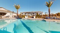 Located at 9975 W Encanto Blvd in Avondale, Arizona, this resort-style outdoor pool area serves as a central amenity space within a residential community. The centerpiece is a generously sized swimming pool with clear, blue-tiled water, accessible via built-in entry steps with handrails, making it welcoming for all users. The surrounding pool deck is finished in a neutral paver material that extends broadly around the pool, providing ample space for lounging and circulation. Rows of blue chaise lounge chairs are arranged along the pool's perimeter, offering residents dedicated sunbathing areas. Tall, mature palm trees are strategically placed throughout the space, lending a tropical ambiance and natural shade while complementing the Arizona desert setting. A covered pavilion with ceiling fans provides a shaded retreat adjacent to the pool, ideal for relaxing out of the direct sun. Additional seating areas and shaded cabana structures with decorative roofing are positioned along the pool's far edge, enhancing the social atmosphere of the space. Ornamental desert-friendly landscaping borders the deck, adding greenery and color. The entire area is enclosed by a safety fence with a gated entry. Under Arizona's abundant blue skies, this pool area delivers a bright, open, and inviting outdoor experience year-round.