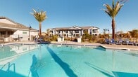 Located at 9975 W Encanto Blvd in Avondale, Arizona, this resort-style outdoor pool area serves as a central amenity space within a residential community. The centerpiece is a generously sized swimming pool with clear, blue-tiled water, accessible via built-in entry steps with handrails, making it welcoming for all users. The surrounding pool deck is finished in a neutral paver material that extends broadly around the pool, providing ample space for lounging and circulation. Rows of blue chaise lounge chairs are arranged along the pool's perimeter, offering residents dedicated sunbathing areas. Tall, mature palm trees are strategically placed throughout the space, lending a tropical ambiance and natural shade while complementing the Arizona desert setting. A covered pavilion with ceiling fans provides a shaded retreat adjacent to the pool, ideal for relaxing out of the direct sun. Additional seating areas and shaded cabana structures with decorative roofing are positioned along the pool's far edge, enhancing the social atmosphere of the space. Ornamental desert-friendly landscaping borders the deck, adding greenery and color. The entire area is enclosed by a safety fence with a gated entry. Under Arizona's abundant blue skies, this pool area delivers a bright, open, and inviting outdoor experience year-round.