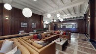 This distinguished hotel lobby showcases a masterful blend of historic craftsmanship and refined interior design. Floor-to-ceiling dark wood paneling lines the walls throughout, lending warmth and gravitas to the space. The coffered ceiling, adorned with intricate plasterwork and crown molding, rises impressively overhead and is illuminated by a series of large, globe-shaped pendant lights that cast a soft, ambient glow across the room.

The flooring is a standout architectural feature — a richly patterned terrazzo surface in deep reds, whites, and blacks arranged in a classic geometric grid, lending an air of historic grandeur. A formal front desk, framed by dark wood millwork and decorative columns, anchors one side of the lobby and includes a traditional key and mail cubby wall, referencing the building's storied past.

The lounge area is generously appointed with large caramel leather sectional sofas and modern accent chairs, accented by an array of colorful throw pillows. Concrete side tables and a textured coffee table add contemporary contrast. A grand staircase with ornate wrought-iron railings and a stained glass window above provides a dramatic focal point near the entrance. Framed historical photographs and greenery complete the welcoming, character-rich atmosphere of this beautifully preserved space.