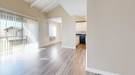 Located at 1919 E Birch St in Brea, California, this one-bedroom, two-bathroom single-floor unit offers a thoughtfully arranged layout that maximizes its compact footprint. The living area immediately impresses with soaring vaulted ceilings accented by exposed wooden beams and a textured ceiling detail, creating a sense of openness well beyond its physical size. Wood-look luxury vinyl flooring runs throughout, lending a clean, cohesive look to every room. Large sliding glass doors and vertical blinds flood the living space with abundant natural light, offering pleasant outdoor views and a bright, airy ambiance. The kitchen features white cabinetry, dark granite-style countertops, stainless steel and black appliances, and recessed lighting. A pass-through counter opens directly to the adjacent dining area, making the space feel connected and functional. A hallway leads to the private bedroom, which includes two closets for ample storage, and connects to both bathrooms. One bathroom features a full tub-shower combination with clean white surround panels and hardwood-style flooring. The neutral warm-beige wall tones throughout the unit complement the modern flooring and create a welcoming, move-in-ready atmosphere.