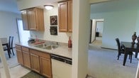 This single-floor residence offers a practical and comfortable layout designed for everyday living. The kitchen is a central highlight, showcasing warm honey-toned wood cabinetry that extends both above and below the countertops, providing generous storage throughout. The countertops feature a speckled granite-look finish that adds a classic touch, complemented by a double stainless steel sink and a built-in dishwasher for added convenience. Flush ceiling lighting keeps the kitchen bright and functional. Decorative wall art adds a homey character to the space. The kitchen opens through a wide passageway into the rest of the home, offering a seamless flow into the carpeted hallway and adjacent living areas. The soft, neutral-toned carpet extends throughout the connecting rooms, creating a cohesive and comfortable feel underfoot. The hallway leads further into the home, hinting at additional rooms beyond, with consistent warm-toned walls and trim framing each space. The overall ambiance is welcoming and practical, with a traditional design aesthetic that emphasizes functionality and ease of movement throughout the single-level floor plan.