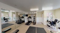 This fitness center is a thoughtfully designed, single-floor exercise facility offering a comprehensive range of workout options. The space is organized into distinct functional zones, providing ample room for both cardio and strength training activities.

The cardio area is equipped with multiple treadmills, an elliptical trainer, and a stationary bike, catering to a variety of aerobic workout preferences. A wall-mounted television is positioned nearby for entertainment during workouts. Full-length mirror panels line one wall, enhancing the sense of openness while allowing users to monitor their form.

The strength training section features a multi-function cable and weight machine along with a leg extension/curl bench, offering a solid range of resistance exercises. Rubber flooring mats are strategically placed throughout the space to provide safe, cushioned surfaces for floor-based workouts and stretching.

Natural light pours in generously through multiple large windows fitted with blinds, offering views of the surrounding greenery and brick exterior. The entrance features a glass-paneled door, further contributing to the bright, open atmosphere. Overhead fluorescent lighting ensures the space remains well-lit at all times.

The interior finishes include light-colored walls with white wainscoting, a textured ceiling, and neutral flooring, creating a clean and functional environment. Safety and informational signage is posted near the equipment area, reflecting a commitment to user well-being.