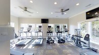 This fitness center is designed to offer a comprehensive and comfortable workout experience within a bright, well-appointed space. The room is anchored by a row of cardio equipment, including treadmills, elliptical machines, and a stationary bike, each outfitted with individual display screens for an engaging exercise session. Additional strength and resistance equipment lines the walls, providing a well-rounded selection of fitness options. Large white-framed windows span the length of one wall, inviting generous amounts of natural light and offering views of the outdoors, contributing to an open and motivating atmosphere. The neutral-toned walls and patterned carpet flooring give the space a clean, polished look. Recessed lighting and dual ceiling fans mounted to the white ceiling ensure the room remains well-lit and comfortably ventilated during workouts. A wall-mounted flat-screen television is positioned prominently above the windows, allowing members to stay entertained while exercising. The overall ambiance is bright, functional, and welcoming, making it a practical and pleasant environment for fitness enthusiasts of all levels.
