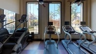 This fitness center is designed to accommodate a comprehensive workout experience within a single, generously proportioned space. The room is distinguished by soaring ceilings fitted with multiple ceiling fans, promoting air circulation throughout. A checkerboard-patterned floor in warm earth tones of brown and tan visually anchors the space and defines workout zones.

The strength training area is well-stocked with a full rack of dumbbells, multiple cable and weight machines, adjustable benches, and a dedicated free-weight zone. Floor-to-ceiling mirrors line portions of the walls, enhancing the sense of openness and allowing users to monitor their form. Stability balls and additional accessories are available for varied training routines.

The cardio section is positioned along a wall of large, floor-to-ceiling windows that frame outdoor views of surrounding greenery and architecture, allowing natural light to flood the area. Treadmills, stair climbers, and cycling equipment are arranged to take full advantage of these views. Wall-mounted televisions provide additional entertainment options. Recessed lighting and pendant-style ceiling fixtures ensure the space remains well-lit throughout the day, creating an inviting and functional fitness environment overall.