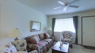 This single-floor space offers a warm and inviting atmosphere, centered around a thoughtfully arranged living room. The room is bathed in natural light streaming through a wide window dressed with elegant drapes and white blinds, creating a bright and airy feel. A ceiling fan with integrated lighting fixture sits overhead, providing both comfort and illumination throughout the space.

The living room is furnished with a classic striped sofa adorned with coordinating throw pillows, complemented by a floral-upholstered wingback chair that adds a touch of traditional charm. A polished dark wood coffee table serves as the centerpiece, accented with decorative candlestick holders. A table lamp on a side table adds warm ambient lighting, while a gold-framed mirror mounted on the wall enhances the sense of depth and elegance.

The neutral-toned walls and light carpeted flooring provide a versatile backdrop that ties the room's classic décor together seamlessly. The overall ambiance is calm, comfortable, and classically styled, making it a welcoming retreat. The space is well-proportioned, offering a comfortable and functional layout suited for relaxing or entertaining.