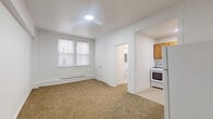Located at 5501 W Washington Blvd in Chicago's West Side, this two-bedroom, one-bathroom unit offers a practical and straightforward layout across a single floor. The main living area features warm beige carpet, crisp white walls, and a skylight-style ceiling fixture that brightens the space. A large double window allows natural light to flow in, providing views of the surrounding neighborhood streetscape. The kitchen is positioned adjacent to the living area and includes warm-toned wooden cabinetry, a gas range, a stainless steel sink, and tile flooring, offering a compact yet functional cooking space. A full-size refrigerator is also included. The bathroom is finished with large-format tan wall tiles surrounding a full bathtub with shower, complemented by a white vanity and tile flooring. The generously sized primary bedroom features plush carpeting and its own dedicated closet with built-in shelving, while the second bedroom is equally well-appointed with carpeted floors and clean white walls. A hallway connects the rooms efficiently, and a dedicated laundry area and two closets add practical storage throughout. Baseboard heating is present in the main living area, ensuring year-round comfort.