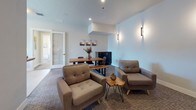 This thoughtfully designed single-floor amenity space offers a welcoming environment suited for both socializing and relaxation. The layout flows seamlessly between distinct functional zones, each finished with contemporary details and a cohesive neutral palette.

The kitchen area serves as a central hub, featuring dark cabinetry, white subway tile backsplash, stainless steel appliances including a double oven, microwave, and French door refrigerator. A generously sized island with a light granite countertop and bar seating provides an inviting space for casual gatherings. A large window fills the kitchen with natural light and views of the surrounding area.

Adjacent to the kitchen, the main lounge area offers a comfortable arrangement of upholstered armchairs and a chaise, paired with live-edge wood accent tables. Wall-mounted sconces provide warm ambient lighting, and plush carpet underfoot adds to the relaxed atmosphere. A dining table with bench seating and a sideboard complete this versatile gathering space.

A dedicated media nook is furnished with plush blue lounge chairs arranged around a wall-mounted flat-screen TV, with built-in speakers and soft sconce lighting creating an intimate viewing experience.

Near the entrance, a reception counter with granite surfaces and bar-height seating offers a convenient welcome area, with an adjacent mail room featuring individual parcel lockers accessible through a glass-panel door.