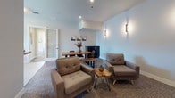 This thoughtfully designed single-floor amenity space offers a welcoming environment suited for both socializing and relaxation. The layout flows seamlessly between distinct functional zones, each finished with contemporary details and a cohesive neutral palette.

The kitchen area serves as a central hub, featuring dark cabinetry, white subway tile backsplash, stainless steel appliances including a double oven, microwave, and French door refrigerator. A generously sized island with a light granite countertop and bar seating provides an inviting space for casual gatherings. A large window fills the kitchen with natural light and views of the surrounding area.

Adjacent to the kitchen, the main lounge area offers a comfortable arrangement of upholstered armchairs and a chaise, paired with live-edge wood accent tables. Wall-mounted sconces provide warm ambient lighting, and plush carpet underfoot adds to the relaxed atmosphere. A dining table with bench seating and a sideboard complete this versatile gathering space.

A dedicated media nook is furnished with plush blue lounge chairs arranged around a wall-mounted flat-screen TV, with built-in speakers and soft sconce lighting creating an intimate viewing experience.

Near the entrance, a reception counter with granite surfaces and bar-height seating offers a convenient welcome area, with an adjacent mail room featuring individual parcel lockers accessible through a glass-panel door.