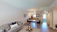 Located at 300-328 S Escondido Blvd in Escondido, Unit #210 is a thoughtfully designed single-floor residence that blends modern aesthetics with everyday functionality. The open-concept layout connects the living area seamlessly to the dining space and kitchen, creating an inviting atmosphere ideal for both relaxing and entertaining. The living area features light-toned walls, warm hardwood-style flooring, and ample natural light that enhances the bright, airy feel of the space. A stylishly furnished dining area sits at the heart of the open floor plan, anchored by a pendant light fixture above a mid-century modern dining table. The kitchen, visible from the living and dining areas, is outfitted with wood-toned cabinetry and a functional layout that keeps essentials within easy reach. Clean white walls and consistent flooring throughout create a cohesive, contemporary look. The unit is part of a well-maintained multi-unit building that features a spacious shared courtyard with seating areas, mature plantings, and upper-level balconies accessible via an exterior walkway. The courtyard benefits from abundant southern California sunshine, offering residents an attractive outdoor gathering space directly outside their doors.