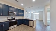 This efficiently designed single-floor unit offers a cohesive, modern aesthetic throughout its thoughtfully arranged rooms. The open-concept kitchen and living area serves as the heart of the space, where bold navy shaker-style cabinetry contrasts sharply against light countertops and white walls. A suite of stainless steel appliances—including a range, over-the-range microwave, dishwasher, and side-by-side refrigerator—equips the kitchen fully. A peninsula with pendant globe lighting overhead defines the transition between the kitchen and living area, which features wood-look plank flooring and a ceiling fan. Natural light filters in through a set of double doors along the living room wall.

The bedroom is a calm, carpeted retreat with a large window fitted with blinds, allowing adjustable natural light. An adjacent walk-in closet provides generous shelving and hanging space along multiple walls. The bathroom is finished to a high standard, featuring navy cabinetry with a dual-sink vanity, framed mirrors with sconce lighting, and a full tub-shower combination with large-format tile surround—all tied together with wood-look flooring consistent with the rest of the unit.