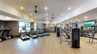 This fitness center offers a comprehensive and well-organized workout environment designed to accommodate a wide range of exercise needs. The space features light hardwood-style flooring throughout, contributing to a clean and modern aesthetic.

Along the windowed wall, a row of treadmills is positioned to take advantage of natural light streaming in from large windows, creating an inviting atmosphere for cardio workouts. Ceiling fans are evenly distributed across the room, ensuring consistent airflow and comfort during exercise sessions.

The strength training area is well-appointed with cable and resistance machines, including recognizable commercial-grade equipment. A decorative accent wall with a bold graphic pattern adds a dynamic visual element to the space, while a mounted flat-screen television provides entertainment during workouts.

Recessed lighting combined with the natural light from the windows ensures the entire space is bright and well-lit at all times. The open layout allows for ease of movement between equipment stations, making the fitness center both functional and user-friendly. Overall, the space strikes a balance between practicality and modern design, offering members a motivating and comfortable environment for their fitness routines.
