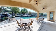 This inviting outdoor amenity space centers around a generously sized swimming pool with crystal-clear blue water, framed by a wide concrete pool deck that provides ample room for lounge chairs and sunbathing. The pool area is complemented by a substantial covered patio structure with an arched, vaulted ceiling finished in smooth stucco and equipped with multiple ceiling fans to ensure comfortable airflow during warm days. The covered space is furnished with round glass-top tables and sling-back chairs, creating a pleasant outdoor dining and gathering environment with direct views of the pool. Stone-accented columns add a refined architectural touch to the covered patio, blending warmth and durability. Large sliding glass doors and screened windows connect the covered patio to an adjacent interior space, allowing for flexible indoor-outdoor flow. The pool deck extends around the full perimeter of the pool, offering additional open-air seating under shade umbrellas. The surrounding landscape features mature trees that provide natural shade and greenery, enhancing the overall ambiance. A wrought-iron perimeter fence encloses the pool area, ensuring privacy and security. The combination of architectural detailing, thoughtful amenities, and lush surroundings creates a comfortable and attractive retreat for outdoor leisure.