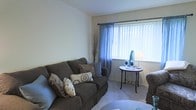 This single-floor residential space offers a comfortable and inviting atmosphere, thoughtfully designed for everyday living. The living room serves as the heart of the space, featuring plush wall-to-wall carpeting that adds warmth underfoot. A large window dressed with elegant blue sheer curtains and vertical blinds allows generous natural light to fill the room, creating a bright and airy ambiance. The seating area is centered around a generously sized, dark upholstered sofa accented with an assortment of decorative throw pillows in complementary patterns and neutral tones. A classic round side table with ornate legs is positioned near the window, topped with a decorative lamp and accent pieces that contribute to the room's polished, homey feel. A matching accent chair rounds out the seating arrangement. The walls are finished in a clean, neutral palette that enhances the sense of openness and provides a versatile backdrop for personal styling. Overall, the space presents a cohesive and functional layout with a calm, understated aesthetic that balances comfort with visual appeal.