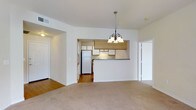 This single-floor residential space offers a well-organized layout designed for comfortable everyday living. The living area is finished with plush wall-to-wall carpeting and painted in crisp white tones, creating a clean and inviting atmosphere. A decorative chandelier with frosted glass shades serves as a central focal point, adding a touch of classic elegance to the space.

The kitchen is accessible via an open archway and features a pass-through counter that connects it to the living area, promoting an open and communicative flow between spaces. The kitchen is equipped with white cabinetry, warm wood-toned upper cabinets, a white refrigerator, and a range with overhead ventilation. The kitchen flooring transitions to hardwood, offering a practical and easy-to-maintain surface.

Arched doorways frame the transitions between rooms, lending architectural character throughout the unit. Multiple doorways are visible from the main living area, suggesting well-distributed access to additional rooms. Neutral wall colors and consistent trim work unify the overall aesthetic, making the space feel cohesive and move-in ready. The layout balances openness with defined areas, offering both functional utility and a comfortable residential ambiance.