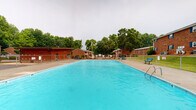 Located at 2205 Woodleaf Rd in Salisbury, North Carolina, this outdoor community pool facility serves as a shared recreational amenity for the surrounding residential complex. The centerpiece of the space is a generously sized rectangular swimming pool filled with clear, vivid blue water, with clearly marked depth indicators along the deck. Multiple entry points via metal handrail ladders provide easy pool access. The surrounding concrete deck offers ample space for poolside activities and is furnished with blue metal benches for seating and relaxation. A wood-clad utility and restroom building anchors one end of the pool area, equipped with restroom facilities, a water fountain, and posted pool rules and safety signage. The pool area is enclosed by a chain-link fence with a gated entry point. The grounds are well-maintained with bordering green lawns and mature trees that provide a natural backdrop, enhancing the open-air atmosphere. The multi-building brick residential complex, featuring blue shutters and exterior stairways, is visible from the pool area, reinforcing its role as a central community gathering space. The facility is designed to accommodate a large number of users and serves as a key outdoor amenity for residents of the community.