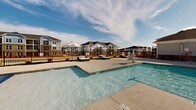 Located at 1055 Winnall Ln in Fayetteville, North Carolina, this outdoor amenity space serves as a premier recreational hub for its residential community. The centerpiece is a generously sized resort-style swimming pool with crystal-clear blue water, complemented by a shallow wading area featuring built-in water fountain elements, making it suitable for a wide range of ages. A dedicated children's splash pad and colorful play structure add a family-friendly dimension to the outdoor experience. The pool deck is finished in a smooth, light-toned concrete surface and is outfitted with rows of lounge chairs and umbrella-shaded seating areas, providing ample space for relaxation under the North Carolina sky. A wrought iron perimeter fence encloses the pool area, ensuring both safety and a defined boundary. A prominently positioned clubhouse anchors one end of the space, featuring a charming architectural design with a covered porch, large arched entry, and a distinctive cupola atop the roofline. The surrounding multi-story residential buildings provide a cohesive neighborhood backdrop. Open skies above and abundant natural light make this an inviting outdoor retreat throughout the day.