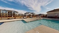 Located at 1055 Winnall Ln in Fayetteville, North Carolina, this outdoor amenity space serves as a premier recreational hub for its residential community. The centerpiece is a generously sized resort-style swimming pool with crystal-clear blue water, complemented by a shallow wading area featuring built-in water fountain elements, making it suitable for a wide range of ages. A dedicated children's splash pad and colorful play structure add a family-friendly dimension to the outdoor experience. The pool deck is finished in a smooth, light-toned concrete surface and is outfitted with rows of lounge chairs and umbrella-shaded seating areas, providing ample space for relaxation under the North Carolina sky. A wrought iron perimeter fence encloses the pool area, ensuring both safety and a defined boundary. A prominently positioned clubhouse anchors one end of the space, featuring a charming architectural design with a covered porch, large arched entry, and a distinctive cupola atop the roofline. The surrounding multi-story residential buildings provide a cohesive neighborhood backdrop. Open skies above and abundant natural light make this an inviting outdoor retreat throughout the day.
