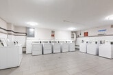 Located at 187 Park St S in Hamilton, Ontario, this shared laundry facility offers a well-organized, functional space designed to serve building residents efficiently. The room is finished with large-format light grey floor tiles that lend a clean, bright aesthetic throughout. The walls feature a two-tone design with white tile on the lower half and a grey accent border, contributing to a tidy and practical interior.

The facility is equipped with multiple top-loading washing machines and front-loading dryers arranged in rows, providing ample capacity for resident use. A stainless steel utility sink is conveniently positioned near the entry, while a built-in laminate folding counter along one wall offers a dedicated surface for sorting and folding laundry. A chair is available at the counter for added convenience. A bulletin board is mounted above the counter for notices and building communications.

Overhead flush-mount lighting ensures the space is well-lit throughout. Safety features include a clearly visible fire extinguisher. Ventilation ducting is visible along one wall, supporting the proper operation of the dryer units. The space is accessed through a utility-style door and maintains a clean, no-frills environment focused entirely on functionality and ease of use for building residents.