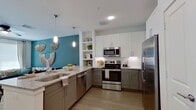 This thoughtfully designed one-bedroom, one-bathroom unit offers a cohesive modern aesthetic across its single floor, unified by light wood-style flooring, crisp white walls, and signature teal accent walls that appear throughout the space.

The open-concept kitchen and living area forms the heart of the unit. The kitchen features two-tone cabinetry — white upper cabinets paired with gray lower cabinets — granite countertops, a central island with seating, stainless steel appliances including a refrigerator, range, and microwave, and built-in open shelving. Pendant lighting illuminates the island, while recessed lighting brightens the full space. The living area benefits from large windows that allow ample natural light.

A bright, white hallway with recessed lighting connects the main living area to the private bedroom, which features a teal accent wall, a ceiling fan, and a generously sized window. The adjacent walk-in closet includes built-in shelving, hanging rods, and dedicated storage space.

The bathroom offers a double-sink vanity with granite countertops, gray cabinetry, a large mirror, and a shower-tub combination, with direct access to the walk-in closet for added convenience.