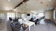 Located in Liberty Lake, Washington, this single-story community clubhouse offers a thoughtfully designed gathering space with a variety of functional areas. The main great room impresses with soaring vaulted ceilings accented by an exposed dark beam, track lighting, and whimsical hanging artwork that adds a playful touch. Large sliding glass doors flood the space with natural light and provide direct access to an outdoor patio, creating a seamless indoor-outdoor connection. The open-concept layout accommodates multiple seating arrangements, a dedicated workstation area, and a dining section suitable for group gatherings. The full kitchen is a standout feature, boasting sleek gray shaker cabinetry, striking marble-look quartz countertops, stainless steel appliances including double wall ovens and a full-size refrigerator, and a spacious island with bar seating. A dedicated media room provides a comfortable, private retreat furnished with plush leather recliners arranged around a wall-mounted flat-screen television with built-in bench seating. A smaller meeting nook with a round table offers a quiet space for intimate conversations. An additional multipurpose room with white cabinetry provides flexible utility. Hardwood-style flooring, neutral tones, and curated artwork throughout contribute to a polished, welcoming ambiance.