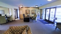 This single-floor space presents a functional and inviting lobby environment designed to serve as a central hub for visitors and staff. Upon entering through the glass-paneled front doors, guests are greeted by a reception area anchored by a large dark wood executive desk, complete with a computer workstation and telephone, accompanied by an ergonomic office chair. A pair of green cushioned accent chairs positioned nearby creates a welcoming seating arrangement for guests. The flooring throughout is carpeted in a muted blue-gray tone, providing a warm and professional atmosphere. Overhead, flush-mount ceiling lights provide consistent, even illumination across the space. The walls are finished in a soft neutral tone and are accented by framed artwork, adding a touch of character to the environment. A patterned sofa is visible in the foreground, contributing to a comfortable waiting area. The lobby also features clear wayfinding signage and a potted plant near the entrance, enhancing the approachable ambiance. Exit signage and additional doorways suggest connectivity to adjoining areas, making this space an efficient and organized point of arrival and departure for the building.