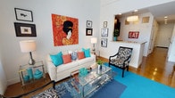 This two-floor space is thoughtfully designed with a bold, eclectic aesthetic that immediately captures attention. The living area features soaring ceilings that amplify the sense of openness, with crisp white walls serving as a neutral backdrop for an eye-catching large-scale artwork — a vivid portrait rendered in warm reds and oranges — that anchors the room. The seating arrangement centers on a cream-toned sofa adorned with teal and coral accent pillows, paired with a striking zebra-print accent chair that adds a playful, fashion-forward element. A glass and chrome coffee table grounds the space while maintaining visual lightness, and a layered rug in rich teal and patterned indigo tones adds depth and warmth to the hardwood flooring beneath. A curated gallery wall featuring framed artwork and photography contributes a personal, artistic dimension to the space. An open pass-through seamlessly connects the living area to the adjacent dining and kitchen zone, where a chandelier pendant light and a decorative "EAT" sign add character. The overall ambiance is lively, stylish, and deliberately curated, combining bold color choices with diverse textures and artistic accents to create a dynamic and inviting interior environment across both floors.