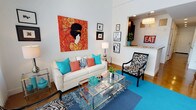 This two-floor space is thoughtfully designed with a bold, eclectic aesthetic that immediately captures attention. The living area features soaring ceilings that amplify the sense of openness, with crisp white walls serving as a neutral backdrop for an eye-catching large-scale artwork — a vivid portrait rendered in warm reds and oranges — that anchors the room. The seating arrangement centers on a cream-toned sofa adorned with teal and coral accent pillows, paired with a striking zebra-print accent chair that adds a playful, fashion-forward element. A glass and chrome coffee table grounds the space while maintaining visual lightness, and a layered rug in rich teal and patterned indigo tones adds depth and warmth to the hardwood flooring beneath. A curated gallery wall featuring framed artwork and photography contributes a personal, artistic dimension to the space. An open pass-through seamlessly connects the living area to the adjacent dining and kitchen zone, where a chandelier pendant light and a decorative "EAT" sign add character. The overall ambiance is lively, stylish, and deliberately curated, combining bold color choices with diverse textures and artistic accents to create a dynamic and inviting interior environment across both floors.