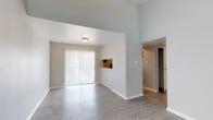 This single-floor residence offers a clean, modern layout designed for comfortable everyday living. The main living area is immediately welcoming, featuring light-toned hardwood-style flooring that runs throughout and lends a cohesive, contemporary feel to the space. Soaring ceilings amplify the sense of openness, making the area feel especially spacious and airy. A large sliding glass door dressed with vertical blinds serves as the focal point of the living room, flooding the interior with generous natural light while providing access to the outdoors. A partial wall with a built-in pass-through window separates the living area from an adjacent space, adding a practical and architectural element to the open floor plan. Neutral, light gray walls complement the pale flooring and contribute to the bright, understated aesthetic throughout. A hallway visible from the main living area leads to additional rooms, suggesting a well-organized and functional layout. The overall ambiance is fresh and move-in ready, with a minimalist design that offers a versatile canvas for personalization. This space combines practical features with an inviting atmosphere, making it a well-rounded single-floor residence.