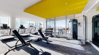 Located at 401 S. Graham Street in Uptown Charlotte, this fitness center offers a generously sized, single-floor workout facility designed with both function and aesthetics in mind. The space makes an immediate visual impact through its bold yellow ceiling panels, which contrast sharply against crisp white walls and recessed lighting, creating a modern, energetic atmosphere throughout.

Floor-to-ceiling glass windows line the perimeter of the facility, flooding the interior with abundant natural light and providing panoramic views of the Charlotte skyline. These expansive windows create a motivating backdrop for workouts while reinforcing the elevated, high-rise setting of the space. A dedicated stretch and functional training area features glass partition walls, hardwood-style flooring, and stability balls, offering a quieter zone for focused training.

The main fitness floor is finished with durable, blue-toned carpet tile and is outfitted with a comprehensive selection of equipment, including treadmills, ellipticals, stationary bikes, and a rowing machine along the window-facing wall, as well as a power rack, adjustable benches, a full dumbbell rack, kettlebells, and a cable machine. Full-length mirrors enhance the sense of space. The overall design balances high-end finishes with a practical, well-organized layout suited for a full range of fitness activities.