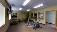 This fitness center offers a dedicated space for exercise and wellness, thoughtfully arranged to accommodate a range of workout routines. The room is finished with durable, textured flooring suited for high-traffic athletic use, and the walls are painted in a neutral yellow-green tone that creates an energizing yet comfortable atmosphere. Overhead, fluorescent lighting ensures the space is brightly and evenly lit throughout. The facility is outfitted with an array of equipment, including a treadmill, an elliptical trainer, a stationary bike, and a multi-station strength training machine, providing users with comprehensive cardio and resistance training options. A weight bench and additional free-weight accessories are also present, further supporting strength-focused workouts. Along one wall, wood-trimmed mirrors reflect the room and help users monitor their form during exercise. A framed informational board near the entrance provides guidance or posted rules for facility users. The space is efficiently organized, making the most of its footprint while maintaining clear pathways between equipment. A security camera is mounted near the ceiling, ensuring a safe and monitored environment. Overall, this fitness center delivers a fully functional and well-maintained workout environment.