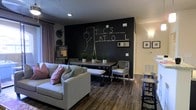 This single-floor space is designed as a welcoming communal lounge that balances comfort with functional versatility. The most striking architectural feature is a full-height chalkboard accent wall painted in deep matte black, which serves as both an artistic focal point and an interactive surface for residents or guests. The contrast between this dark statement wall and the surrounding light-toned walls creates a dynamic visual depth throughout the room. Rich, dark hardwood-style flooring runs throughout the space, grounding the interior with warmth and cohesion. The lounge area is anchored by a plush, light gray sofa accented with colorful throw pillows, offering a relaxed setting for conversation or casual gatherings. Adjacent to the seating area is a sturdy rectangular dining table with woven-back chairs and a matching bench, complemented by small potted greenery that adds a natural, refreshing touch. Recessed lighting provides warm, focused illumination throughout, while sheer curtains frame the windows, softening the overall ambiance. A high-top bar table with stools is also present, adding another layer of seating flexibility. The space is further personalized with framed wall art and a bulletin board, making it feel both curated and community-oriented. Overall, the design is modern, inviting, and highly functional.