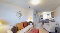 Located at 113 Brownlea Dr in Greenville, NC, this single-floor residence offers a comfortable and functional layout designed for easy living. The living room serves as the heart of the home, featuring plush carpeted flooring, neutral-toned walls, and a textured ceiling that adds subtle character to the space. A generously sized sofa with colorful accent pillows provides a cozy seating area, complemented by warm ambient lighting from a classic table lamp and a ceiling light fixture. Decorative wall art adds a personalized touch to the inviting atmosphere. The open sightline from the living area extends down a hallway that connects seamlessly to the kitchen and dining area, which is visible in the background and features wood-toned cabinetry and countertops. Natural light filters through a window near the kitchen, brightening the interior and enhancing the airy feel of the space. The hallway also leads to additional rooms, suggesting a well-organized floor plan that maximizes functionality. Overall, this home presents a warm, approachable aesthetic with cohesive finishes throughout, making it a comfortable and practical living environment.