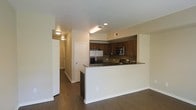 This single-floor residence is designed with an open, functional layout that integrates the kitchen and living areas into one cohesive space. The kitchen is a standout feature, equipped with stainless steel appliances including a French door refrigerator and a built-in microwave and oven combo. Rich, dark wood cabinetry lines the upper and lower walls, providing ample storage, while dark granite countertops add a sleek, modern aesthetic. A peninsula counter extends into the living space, creating a natural divide between the kitchen and the main living area while also offering additional prep and seating space.

The flooring throughout is a dark hardwood-style material that lends warmth and continuity across the open floor plan. Neutral, light-toned walls complement the darker finishes, keeping the space feeling bright and balanced. Recessed lighting and flush-mount ceiling fixtures provide consistent illumination throughout. A hallway visible from the main area leads to additional rooms, suggesting a thoughtful layout that balances open communal areas with more private spaces. The overall ambiance is clean, contemporary, and practical.