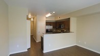 This single-floor residence is designed with an open, functional layout that integrates the kitchen and living areas into one cohesive space. The kitchen is a standout feature, equipped with stainless steel appliances including a French door refrigerator and a built-in microwave and oven combo. Rich, dark wood cabinetry lines the upper and lower walls, providing ample storage, while dark granite countertops add a sleek, modern aesthetic. A peninsula counter extends into the living space, creating a natural divide between the kitchen and the main living area while also offering additional prep and seating space.

The flooring throughout is a dark hardwood-style material that lends warmth and continuity across the open floor plan. Neutral, light-toned walls complement the darker finishes, keeping the space feeling bright and balanced. Recessed lighting and flush-mount ceiling fixtures provide consistent illumination throughout. A hallway visible from the main area leads to additional rooms, suggesting a thoughtful layout that balances open communal areas with more private spaces. The overall ambiance is clean, contemporary, and practical.