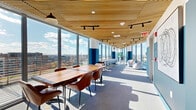 Located on an upper floor at 7 New York Ave NE in Washington, DC, this contemporary collaborative workspace offers an elevated experience defined by thoughtful design and abundant natural light. The space is lined with expansive floor-to-ceiling glass windows along its entire length, framing panoramic views of the Washington, DC skyline and surrounding neighborhoods — a standout architectural feature that fills the interior with daylight throughout the day.

The warm slatted wood ceiling adds a natural, refined aesthetic that contrasts elegantly with the bold teal and white color-blocked walls beneath. Track lighting runs the length of the ceiling, providing flexible, adjustable illumination. Grey carpet flooring grounds the space with a clean, professional finish.

The layout accommodates multiple zones designed for different working and gathering needs. A large live-edge wood conference table with leather chairs anchors one end of the space, ideal for meetings or collaborative work sessions. A window-facing counter bar with upholstered stools runs along the glass wall, offering individual workstations with unobstructed city views. At the far end, a casual lounge area with soft seating provides a relaxed setting for informal conversations. Two hallways connect the zones and lead to additional building amenities. The overall ambiance is bright, modern, and professionally inviting.