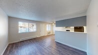 This single-floor unit offers a practical and well-appointed layout designed for comfortable everyday living. The open-concept living area is anchored by durable hardwood-style plank flooring, a textured ceiling, and a ceiling fan, creating an inviting and functional space. A large window along the living room wall allows natural light to fill the room, while a wall-unit air conditioner and baseboard heating provide year-round climate control. A half-wall partition with a blue accent panel separates the living area from the kitchen, adding a subtle design element to the open floor plan.

The galley-style kitchen features gray cabinetry, light countertops, and black appliances including a range and refrigerator, complemented by hardwood-style flooring and overhead fluorescent lighting. The layout maximizes storage and efficiency within a compact footprint.

The bedrooms are carpeted for comfort, each featuring a window with natural light, baseboard heating, and neutral walls. One bedroom includes a built-in closet with shelving. The bathroom is clean and functional, offering a tub-shower combination with white tile surround, hardwood-style flooring, and baseboard heating. Throughout the unit, a consistent neutral color palette and updated finishes contribute to a tidy, move-in-ready atmosphere.