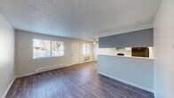 This single-floor unit offers a practical and well-appointed layout designed for comfortable everyday living. The open-concept living area is anchored by durable hardwood-style plank flooring, a textured ceiling, and a ceiling fan, creating an inviting and functional space. A large window along the living room wall allows natural light to fill the room, while a wall-unit air conditioner and baseboard heating provide year-round climate control. A half-wall partition with a blue accent panel separates the living area from the kitchen, adding a subtle design element to the open floor plan.

The galley-style kitchen features gray cabinetry, light countertops, and black appliances including a range and refrigerator, complemented by hardwood-style flooring and overhead fluorescent lighting. The layout maximizes storage and efficiency within a compact footprint.

The bedrooms are carpeted for comfort, each featuring a window with natural light, baseboard heating, and neutral walls. One bedroom includes a built-in closet with shelving. The bathroom is clean and functional, offering a tub-shower combination with white tile surround, hardwood-style flooring, and baseboard heating. Throughout the unit, a consistent neutral color palette and updated finishes contribute to a tidy, move-in-ready atmosphere.